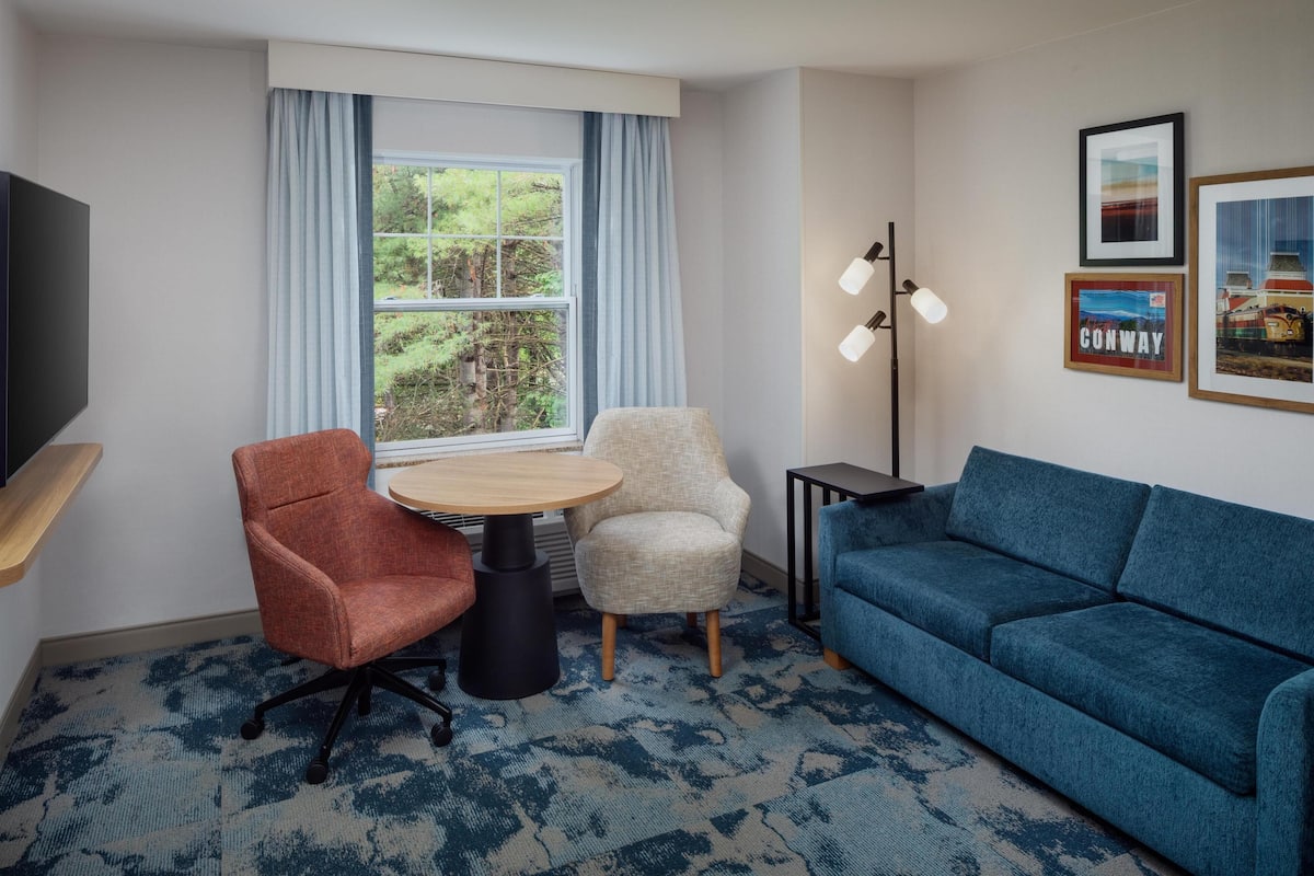 A room with a blue sofa, a red chair, a beige armchair, a round table, and a TV on a shelf.