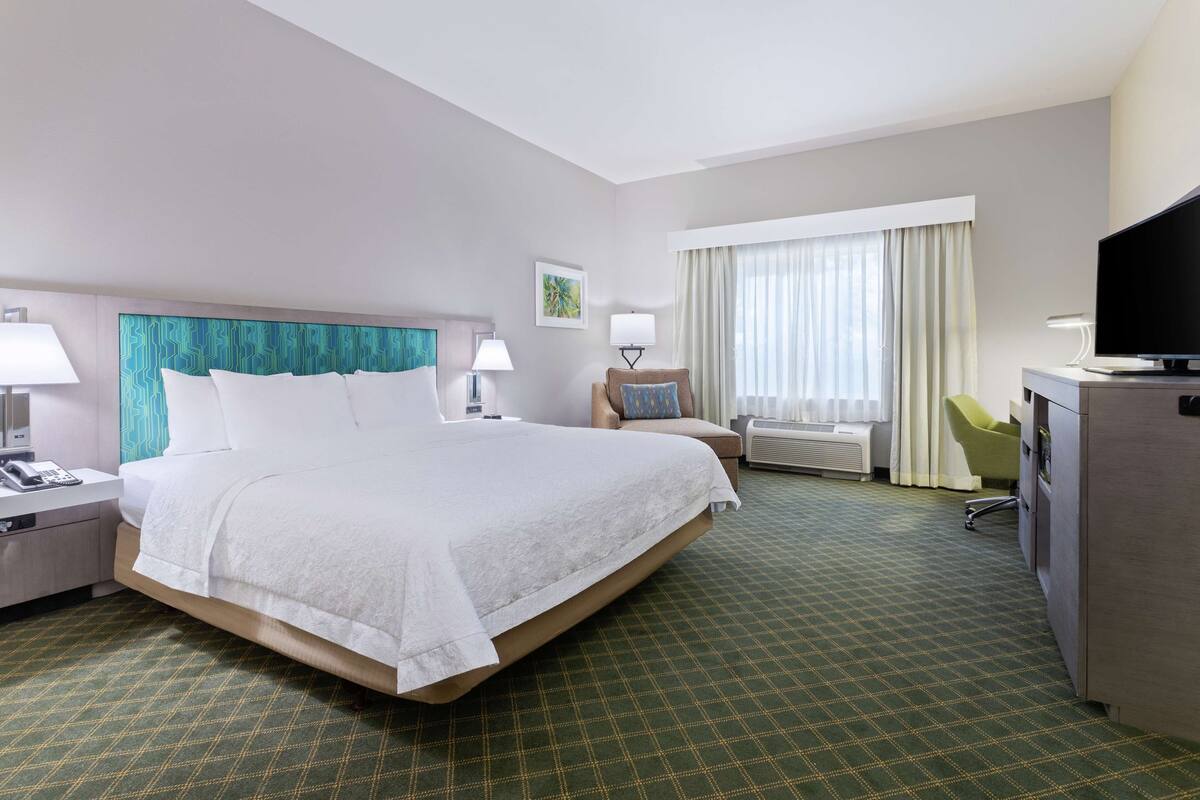 Habitación de hotel con una cama grande, un escritorio, una silla, un televisor y una ventana con cortinas.