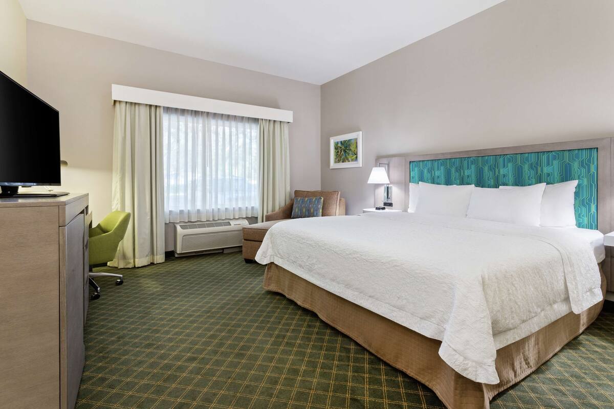 Una habitación de hotel con una cama grande, un escritorio con televisor de pantalla plana, una silla, una ventana con cortinas y una lámpara fijada a la pared.