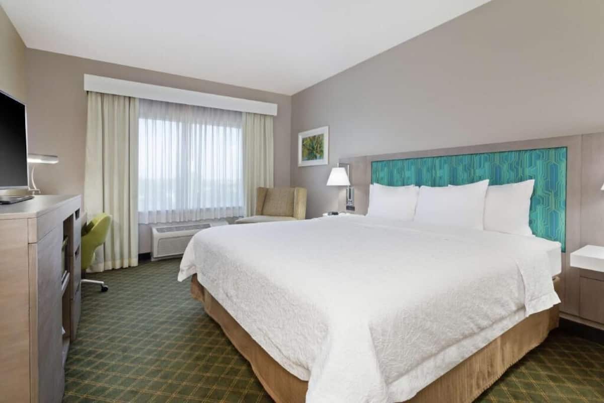 Habitación de hotel con una cama grande, un escritorio con computadora, una silla, una ventana con cortinas y un televisor montado en la pared.
