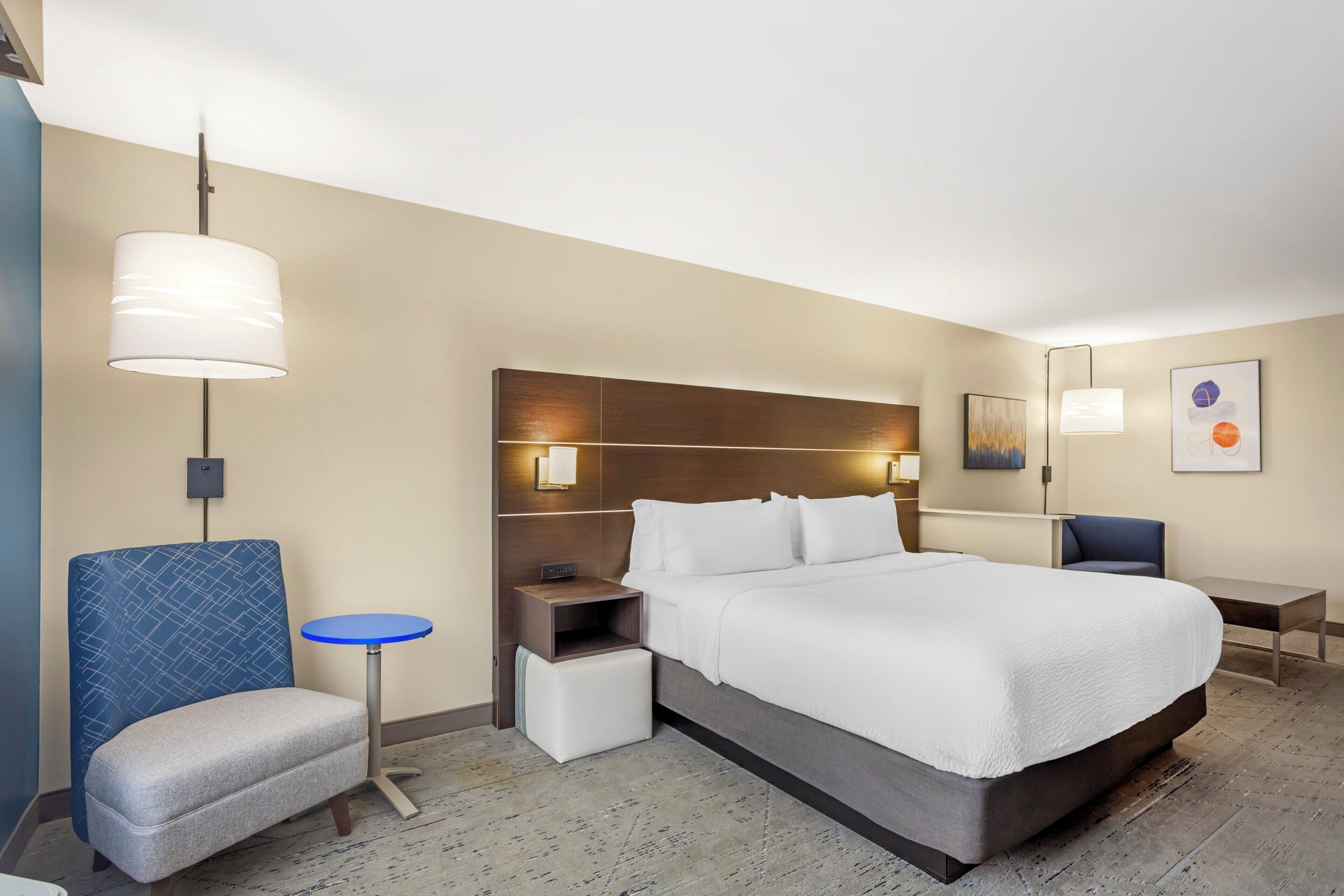 Una habitación de hotel con una cama grande, una mesita de noche, una silla, una pequeña mesa y una lámpara de pared.