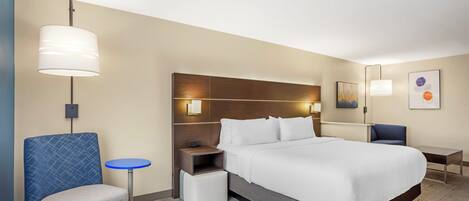 Una habitación de hotel con una cama grande, una mesita de noche, una silla, una pequeña mesa y una lámpara de pared.