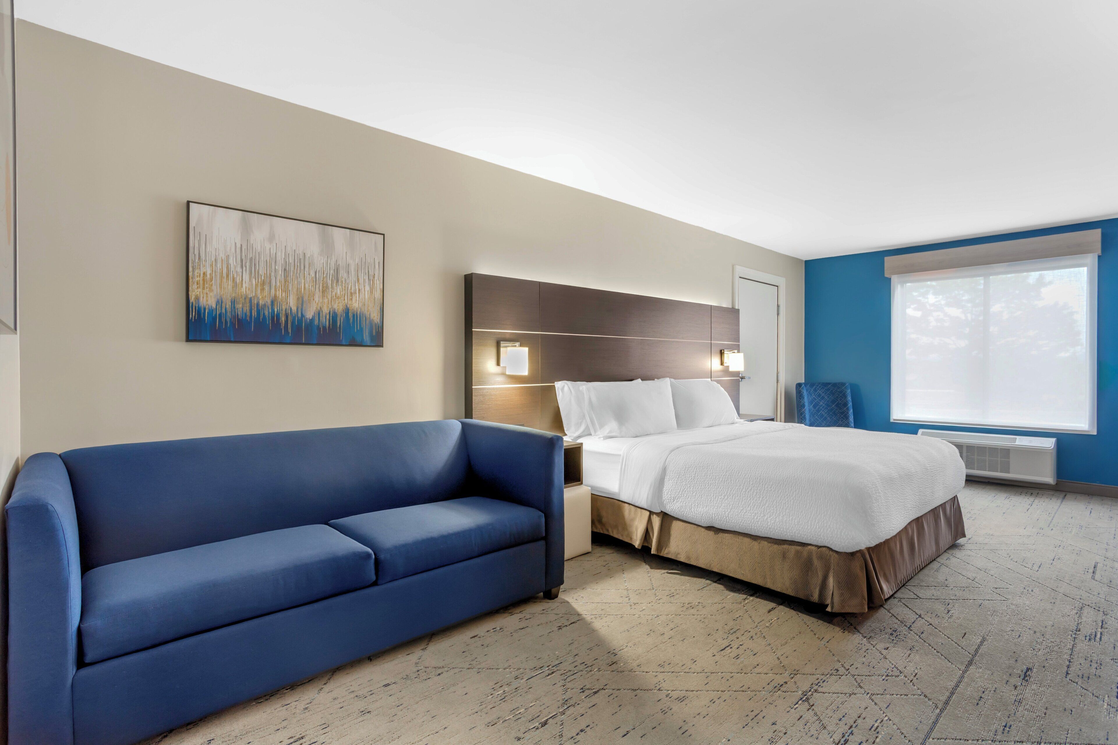 Une chambre d’hôtel avec un lit, un canapé bleu, une table de chevet, une lampe fixée au mur et une grande fenêtre.