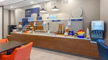 A modern breakfast area with a counter displaying various food options, a coffee machine, and seating arrangements.