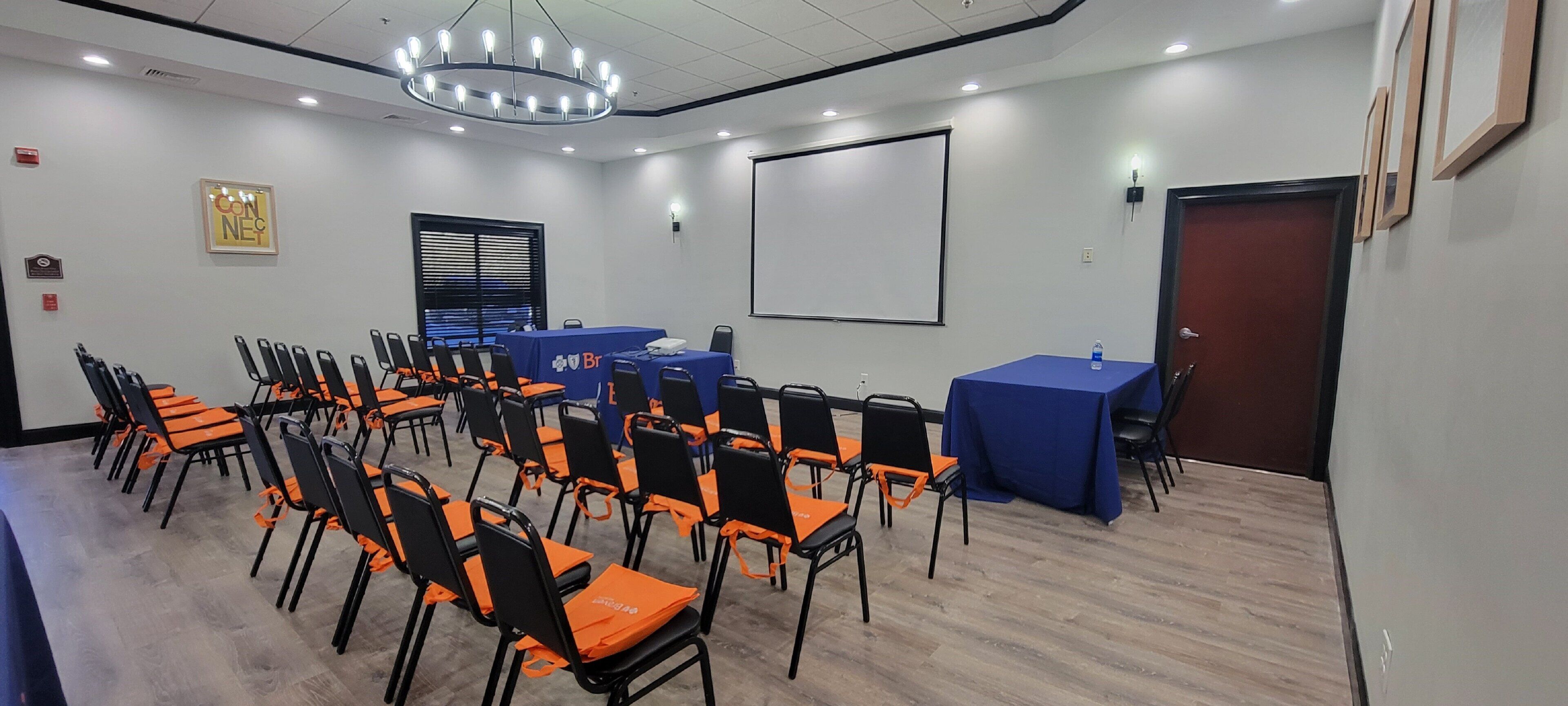 A conference room with rows of orange and black chairs, a blue table, a projector screen, and a door.