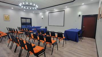 A conference room with rows of orange and black chairs, a blue table, a projector screen, and a door.