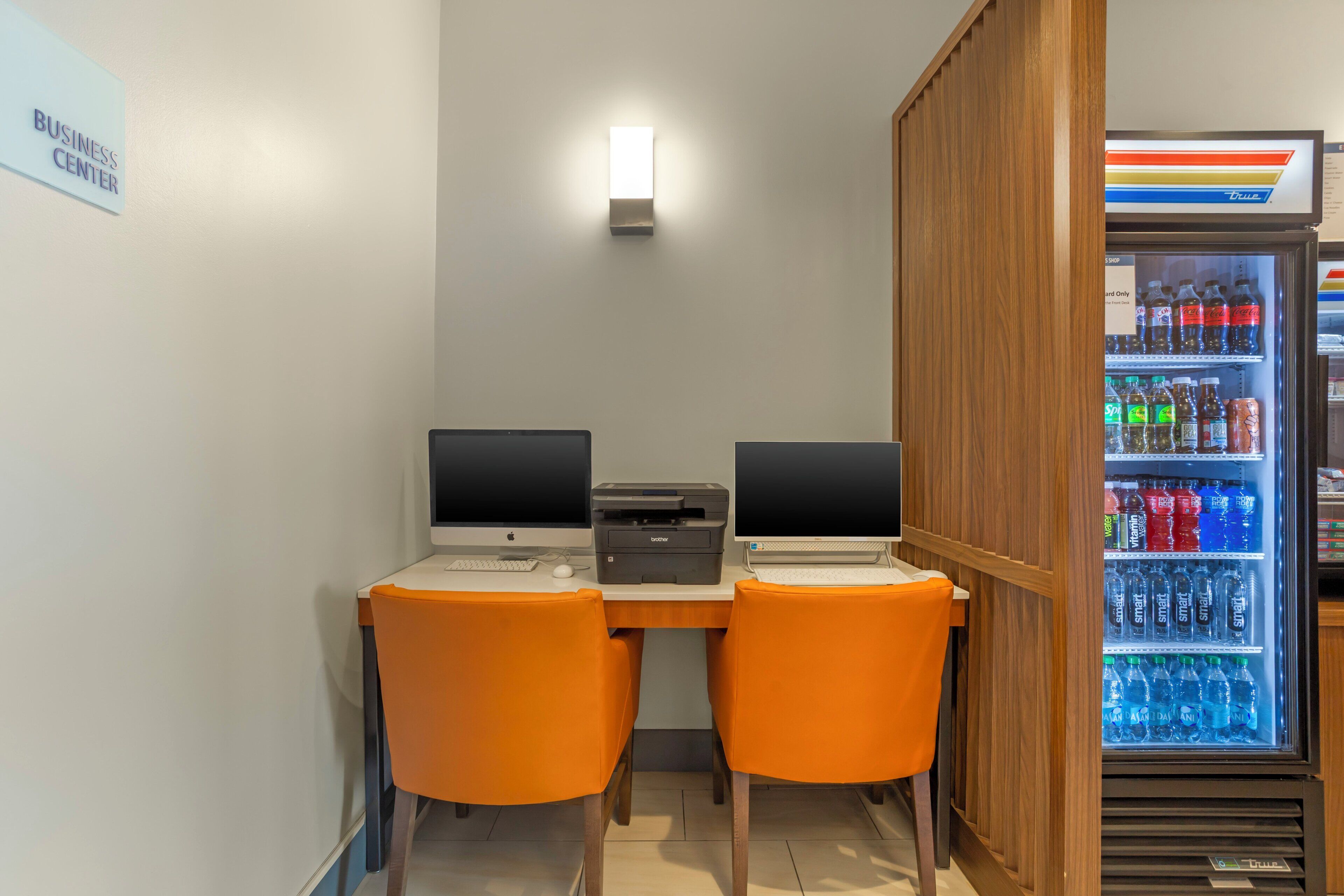 A workspace with two computers, a printer, and a refrigerator stocked with beverages.