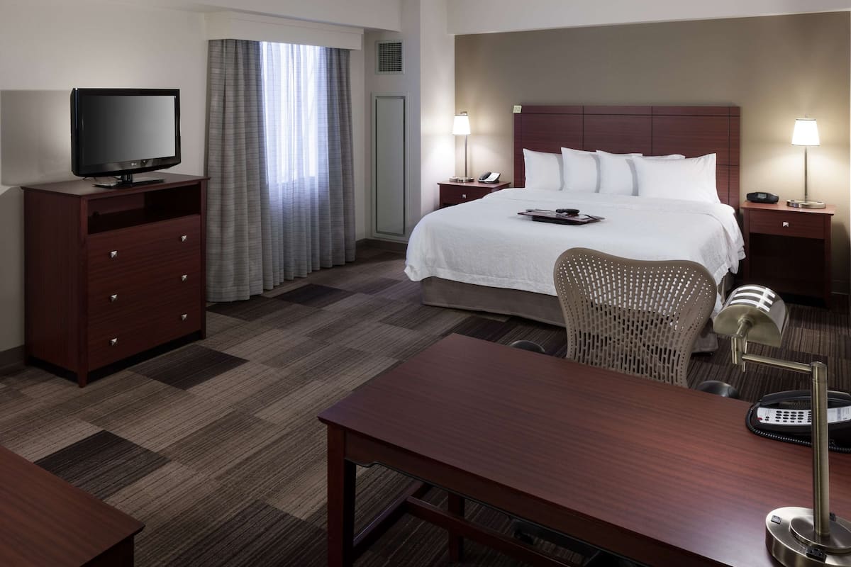 A hotel room with a large bed, a desk, a chair, a TV, and a lamp.