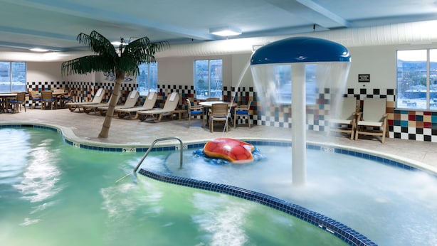 An indoor swimming pool with lounge chairs and a palm tree, a water slide, and a view of the city through large windows.