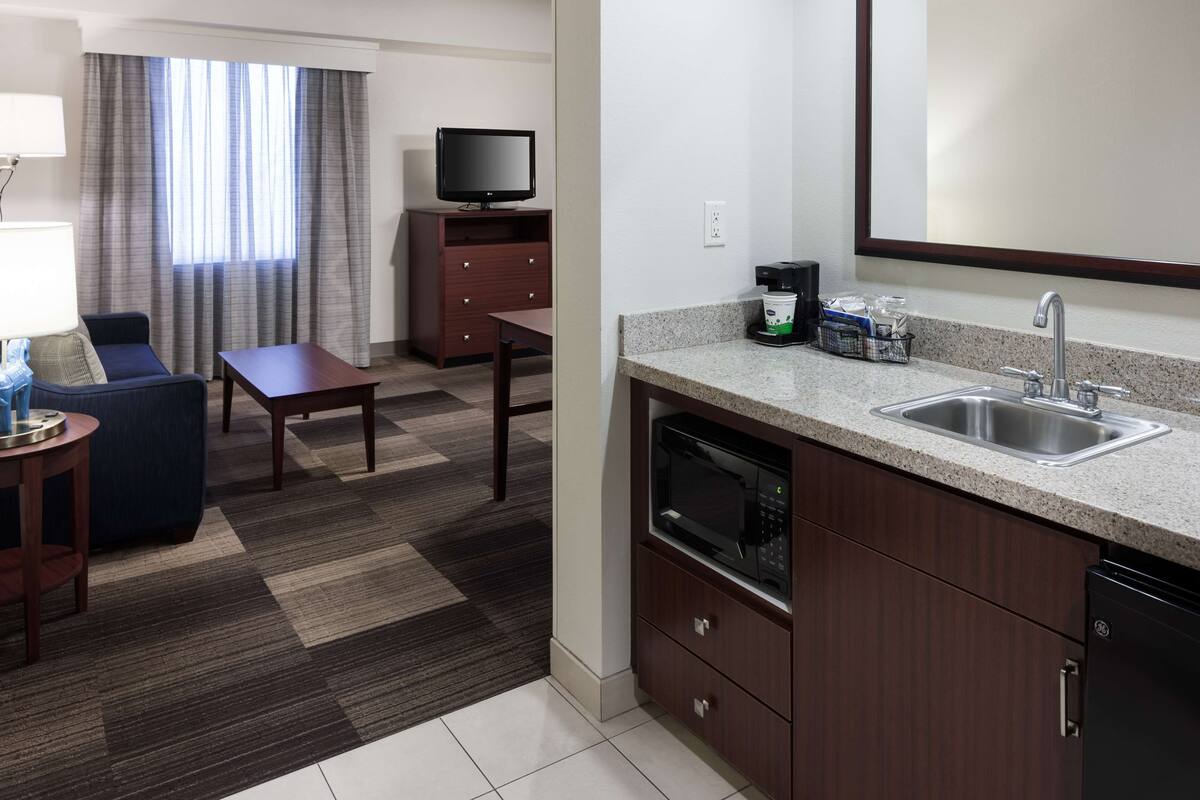 A compact hotel room with a kitchenette, a flat-screen TV, a dining area with a table and chairs, and a sofa.