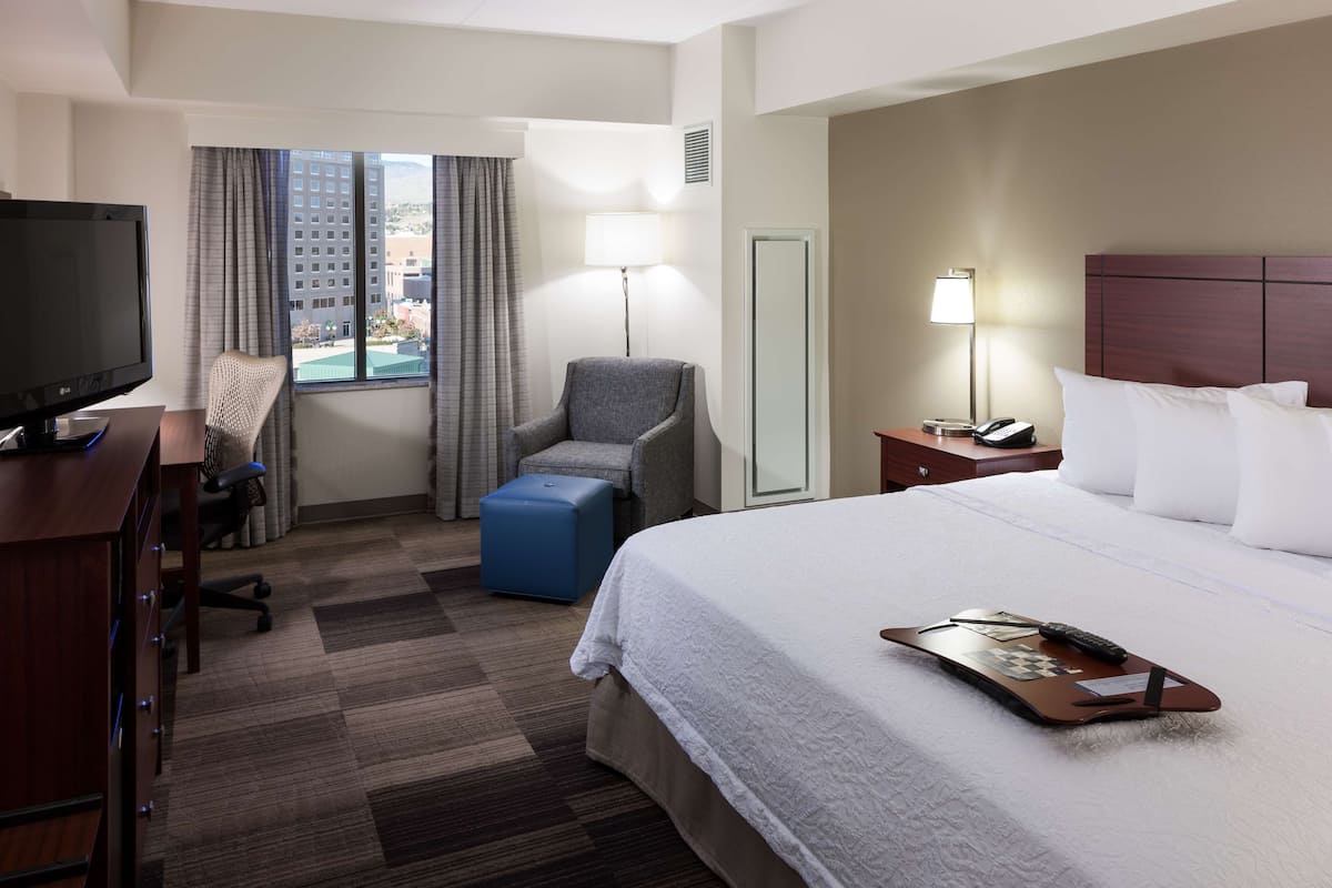 A hotel room with a bed, a desk with a TV, a chair, a lamp, and a view of buildings outside.