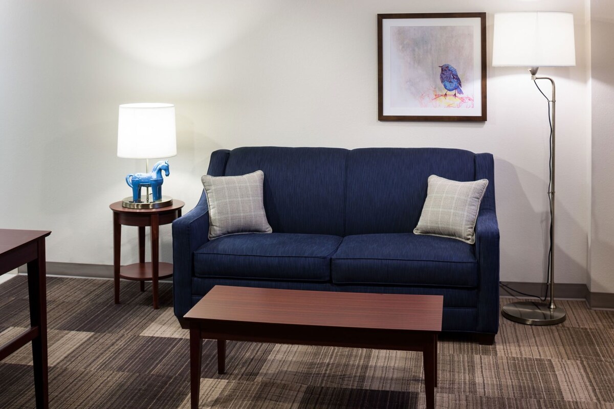 A room with a blue sofa, two pillows, a wooden coffee table, a lamp, a framed picture, and a floor lamp.