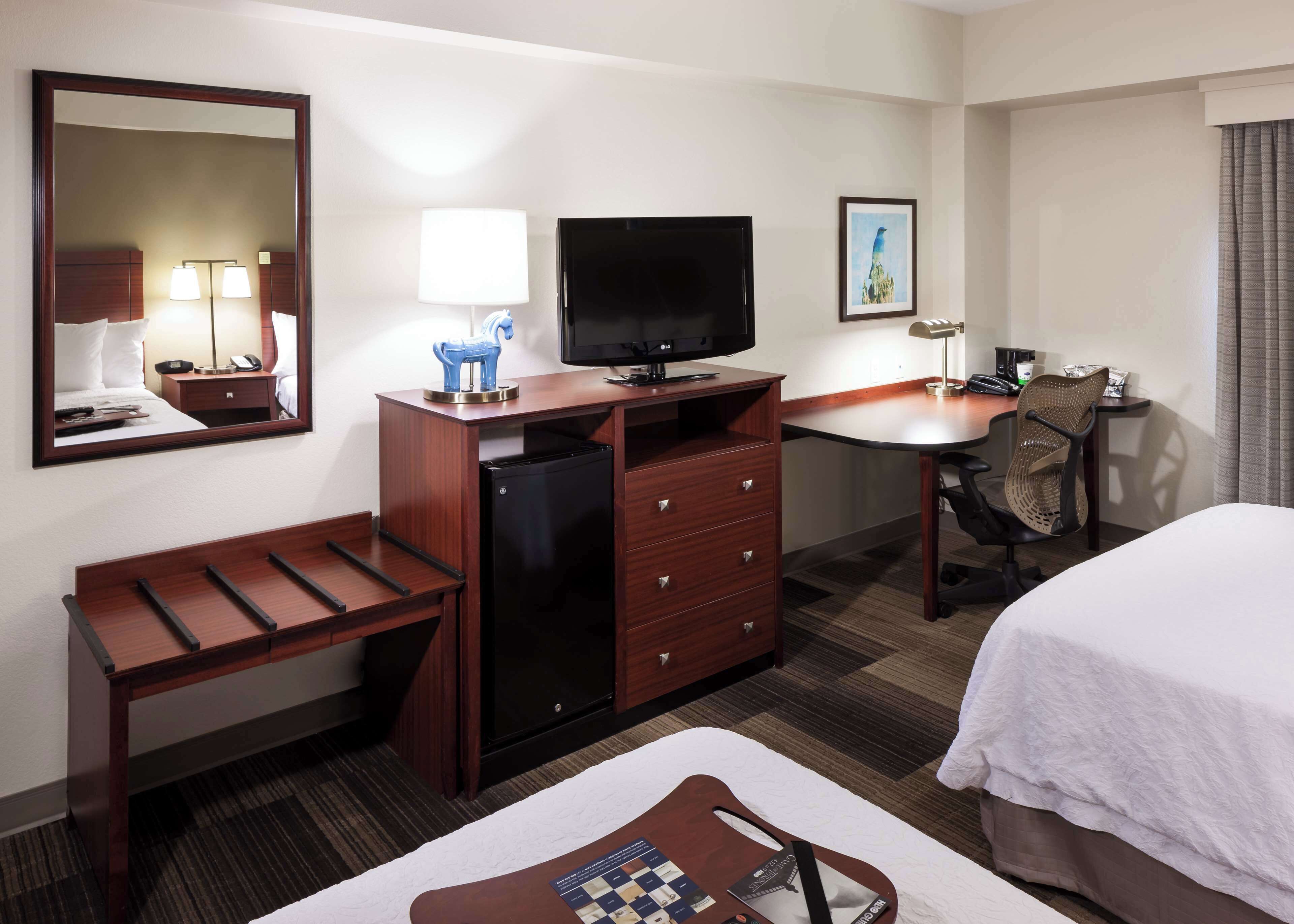 A hotel room with a bed, desk, chair, TV, refrigerator, and a mirror.