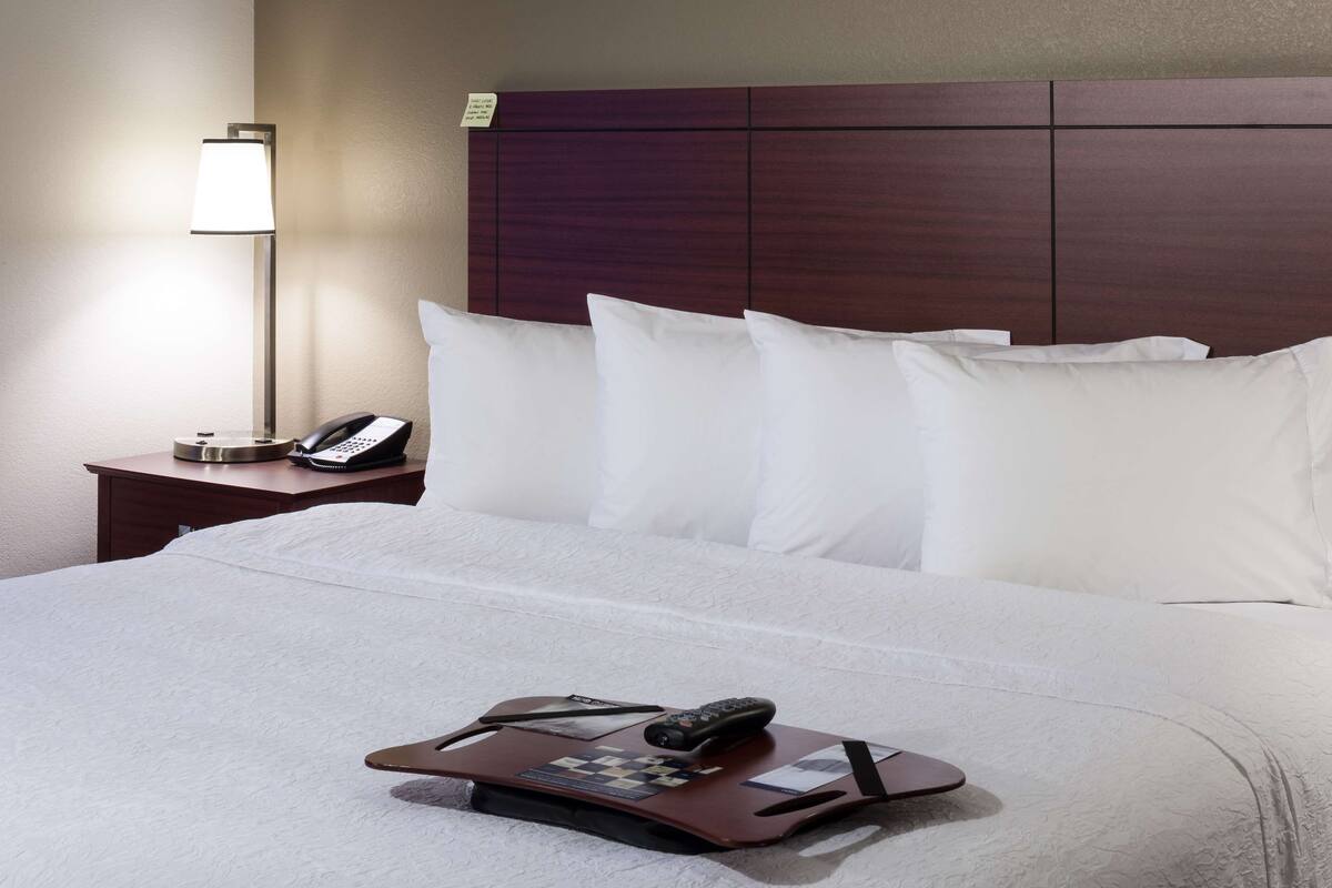 A hotel room with a neatly made bed, a bedside lamp, a phone, and a tray with a remote control and a card.