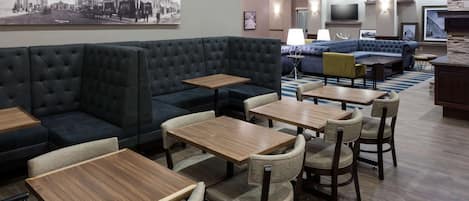 A modern hotel lobby with a large dark sofa, multiple wooden tables, and a seating area with a blue couch and yellow chairs.