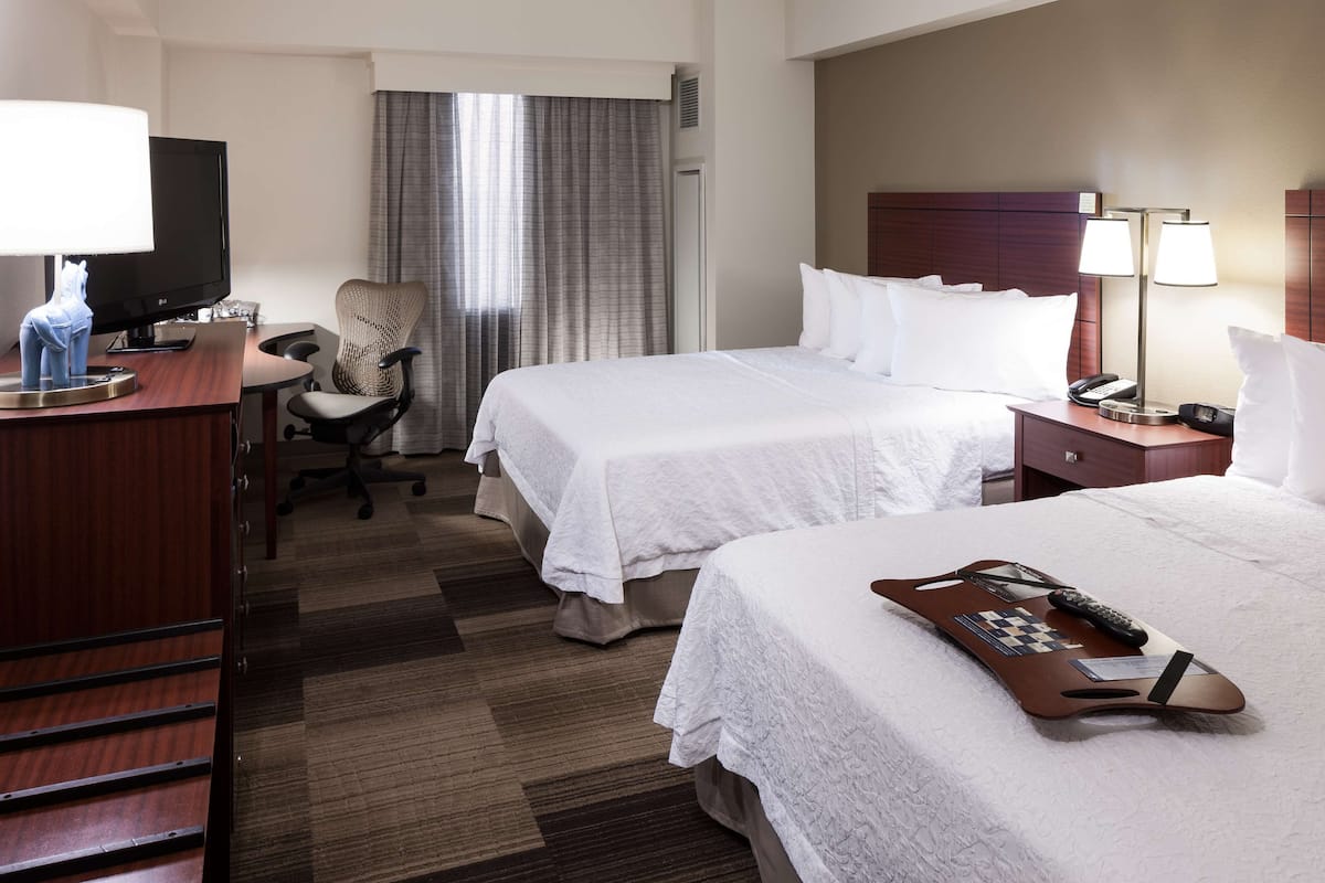 A hotel room with two beds, a desk with a computer, a chair, a TV, and a lamp.