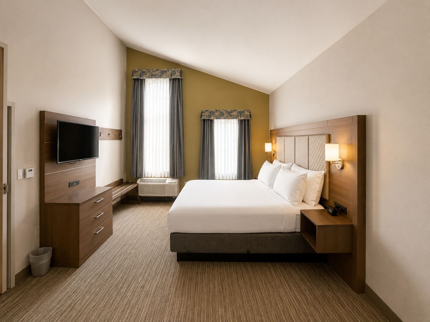 Una camera d'albergo con un letto, una televisione, un comodino con lampada, una cassettiera e una finestra con tende.
