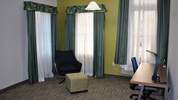 A room with a desk, chair, and a small ottoman. There are two windows with curtains, a lamp, and a wall socket.