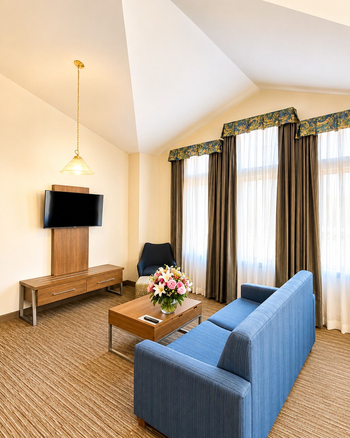 A hotel room with a sofa, a TV mounted on a wooden cabinet, a coffee table with a vase of flowers, and a chair.