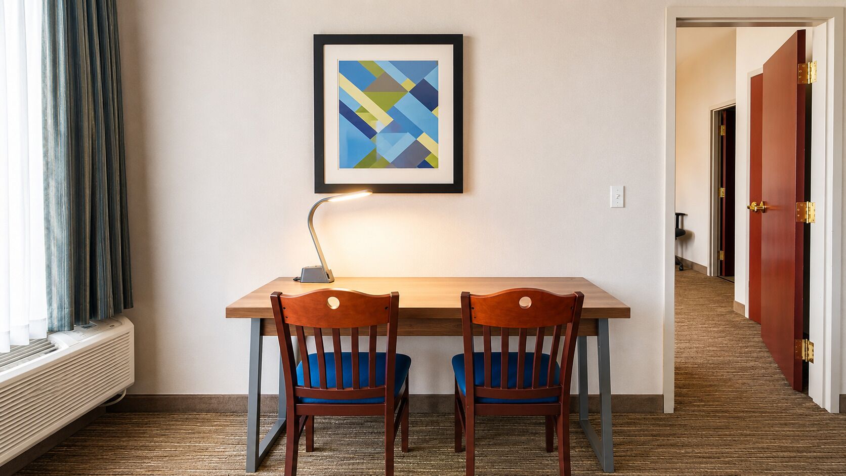 A hotel room with a wooden desk, two chairs, a framed abstract painting, a standing lamp, and a radiator.