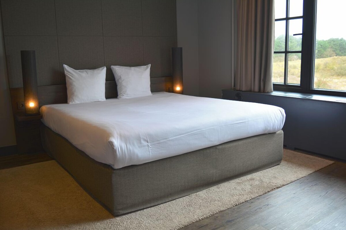 A large bed with white linens in a modern bedroom with a view of the outdoors through a window.
