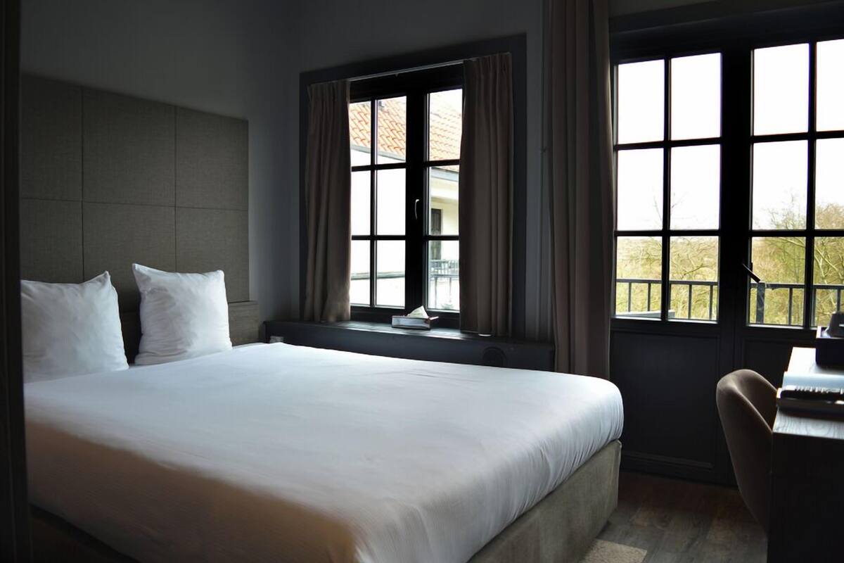 A hotel room with a large bed, a desk, and two windows offering views of the outdoors.
