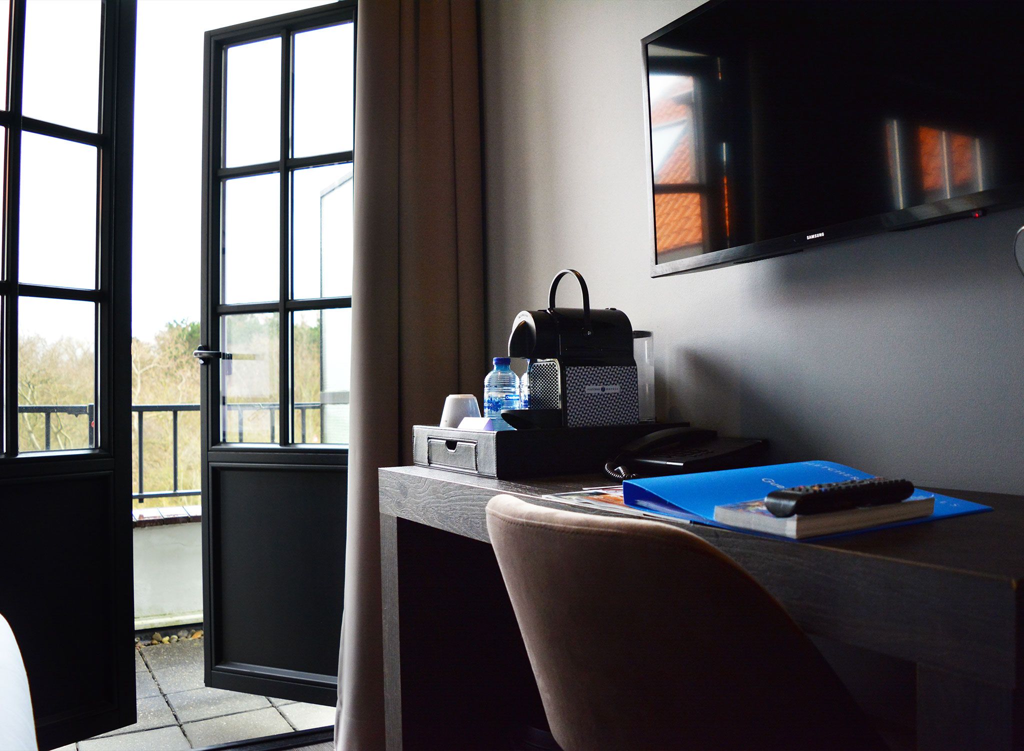 A hotel room with a desk, a chair, a television, and a window with a view of trees.