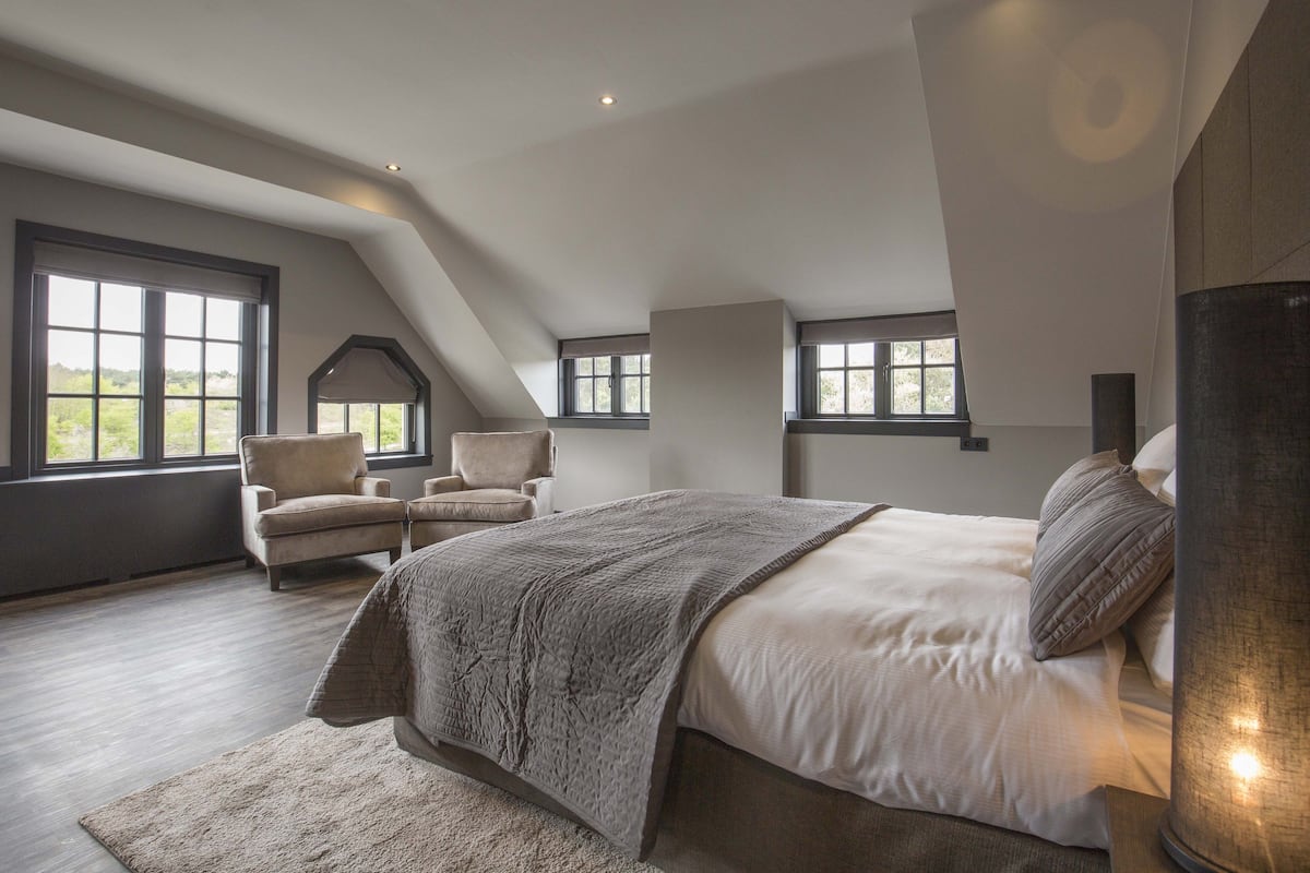A bedroom with a large bed, two armchairs, a window with a view, and a ceiling with recessed lighting.