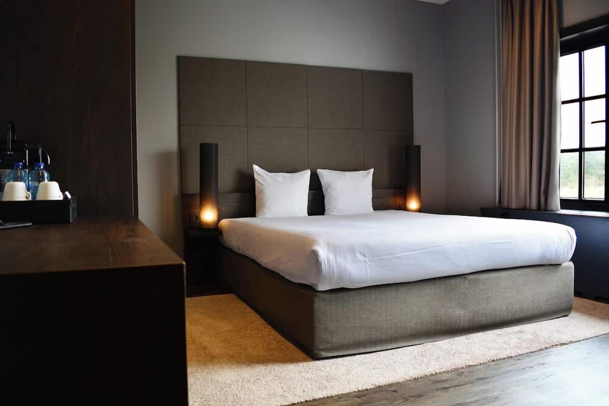 A modern bedroom with a large bed, a dark headboard, bedside lamps, and a coffee station.