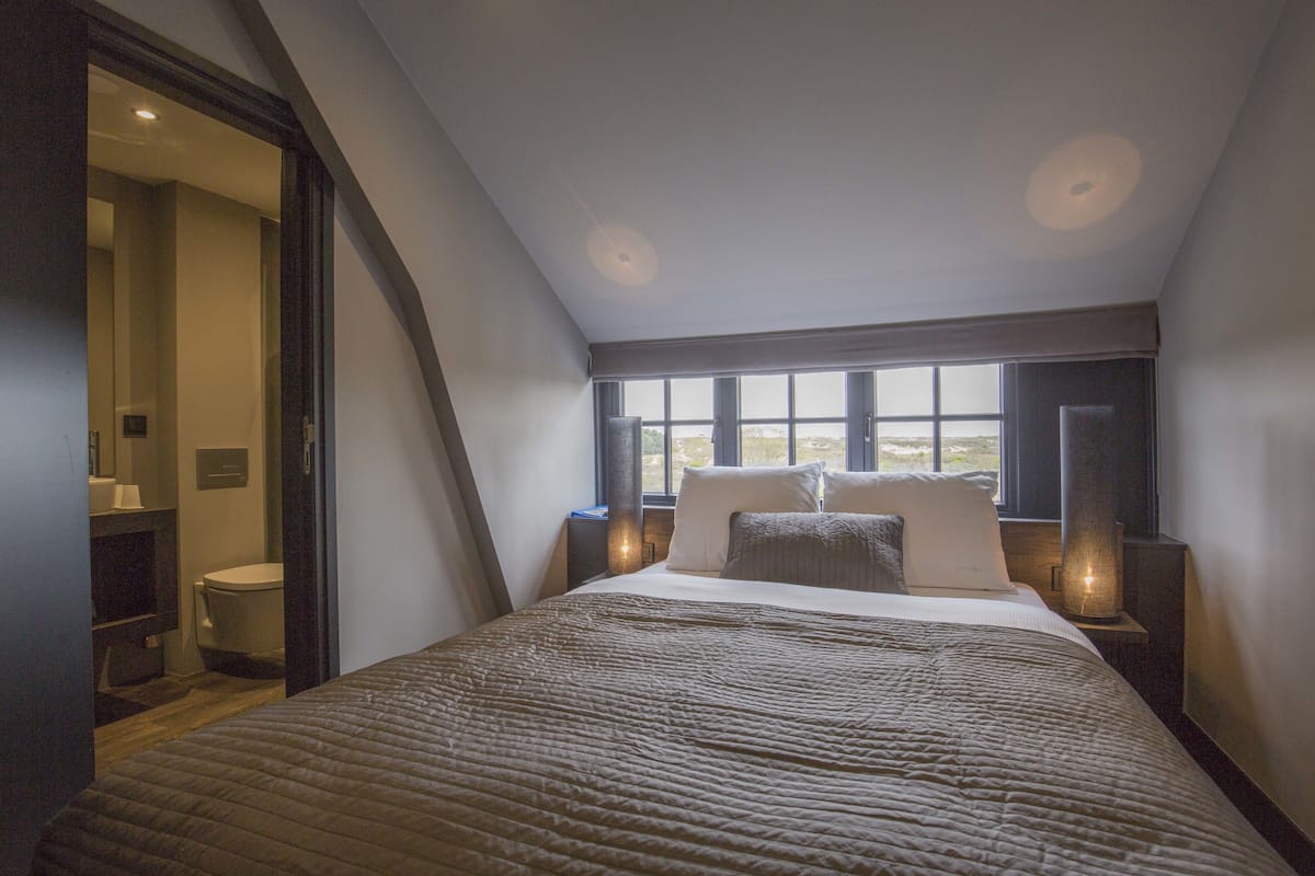 A bedroom with a large bed, a window with a view, and a nightstand with a lamp.