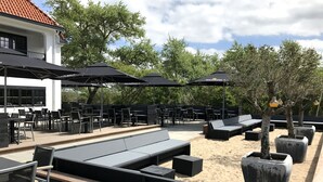 An outdoor seating area with tables and chairs, umbrellas, and a sand area with lounge beds. - Fletcher Boutique Hotel Duinoord (Wassenaar)