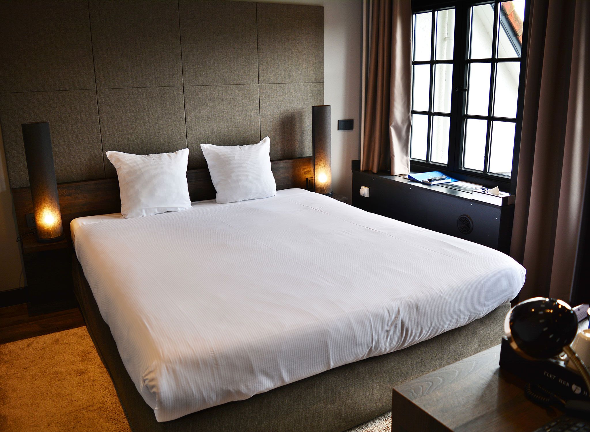 Quarto de hotel com uma cama grande, duas luminárias de cabeceira, mesa de cabeceira, janela com cortinas e uma escrivaninha com telefone.