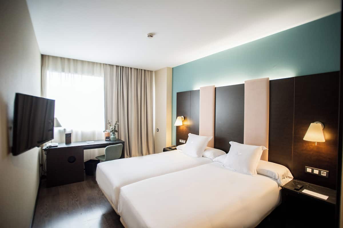 A hotel room with two beds, a desk, a TV, and a window with curtains.
