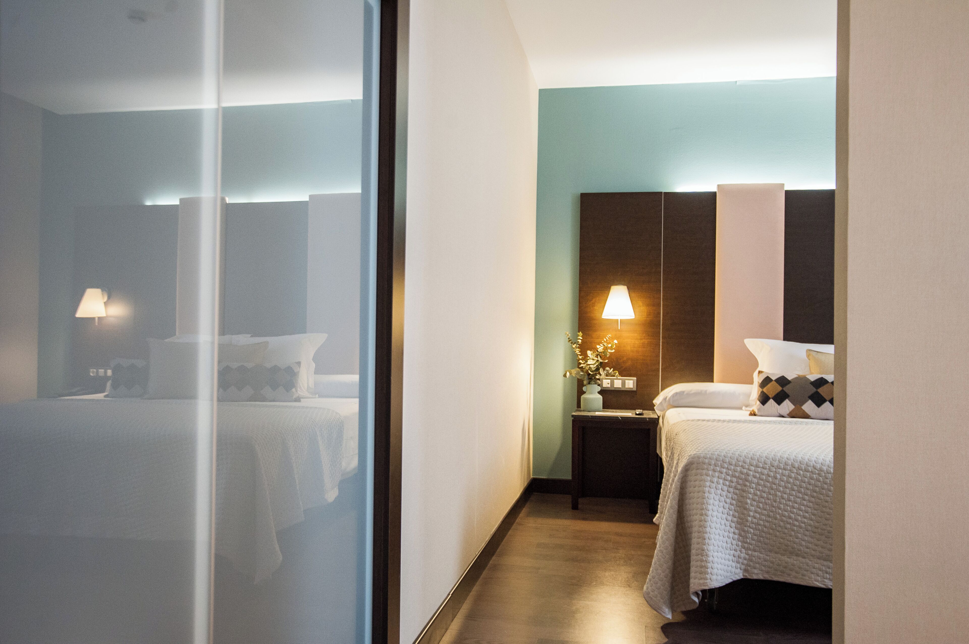 A hotel room with two beds, a nightstand with a lamp, and a wall-mounted light fixture.