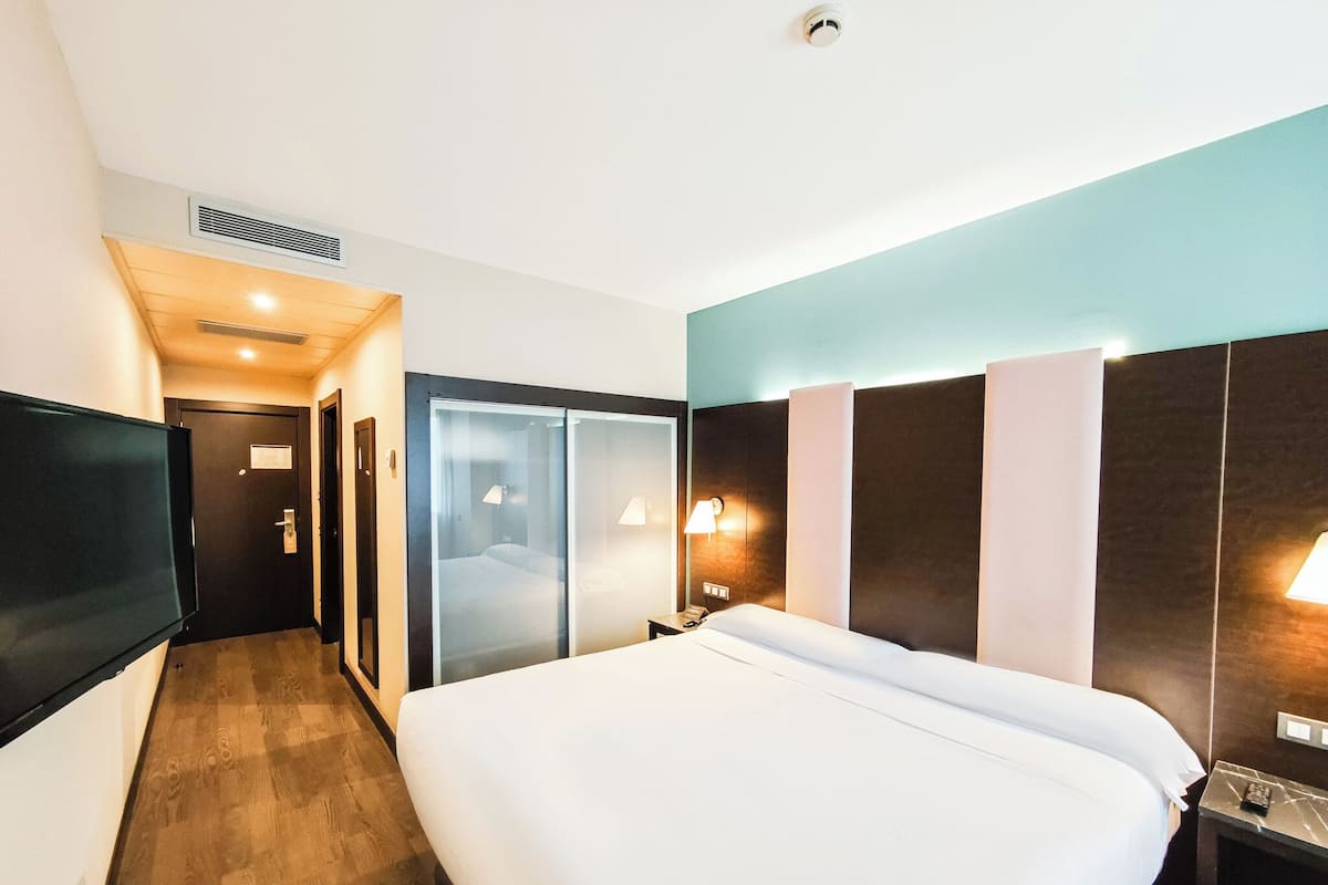 A hotel room with a large bed, a TV mounted on the wall, a bedside table with a lamp, and a door leading to a bathroom.