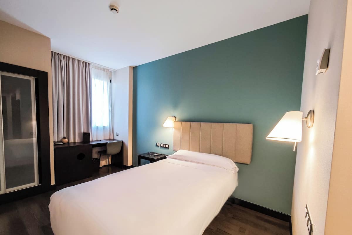 A hotel room with a large bed, a desk, a chair, and a wall-mounted lamp.