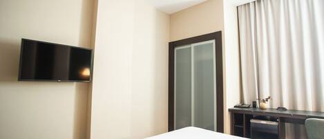 A hotel room with a bed, a desk with a computer, a TV mounted on the wall, and a sliding door.
