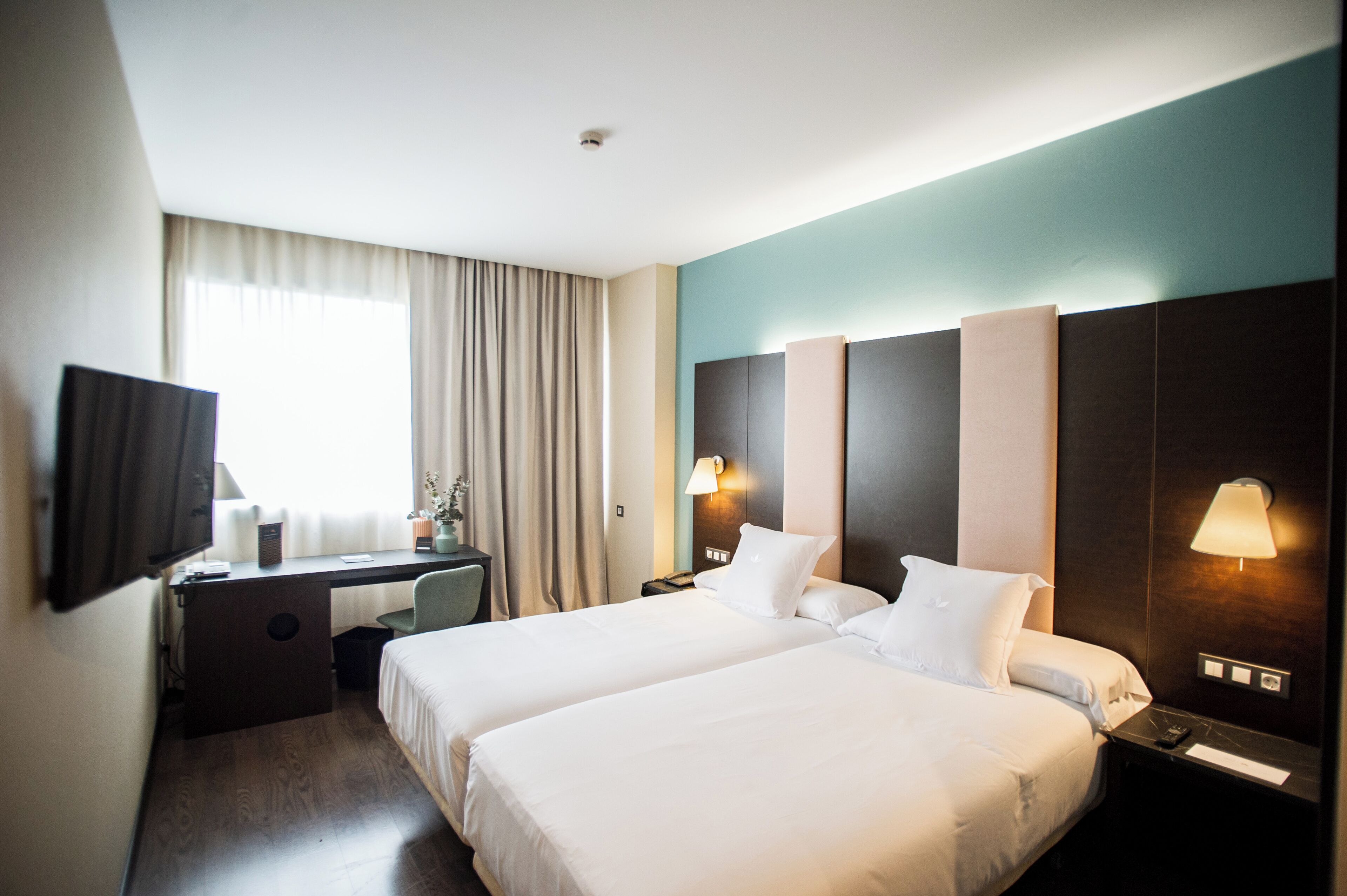 A hotel room with two beds, a desk, a TV, and a window with curtains.
