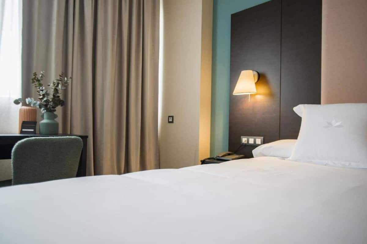 A hotel room with a bed, a desk with a lamp, a vase with flowers, and a window with curtains.