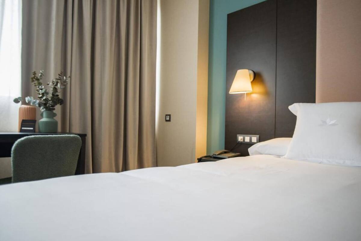 Een hotelkamer met een bed, een bureau met lamp, een vaas met bloemen en een raam met gordijnen.