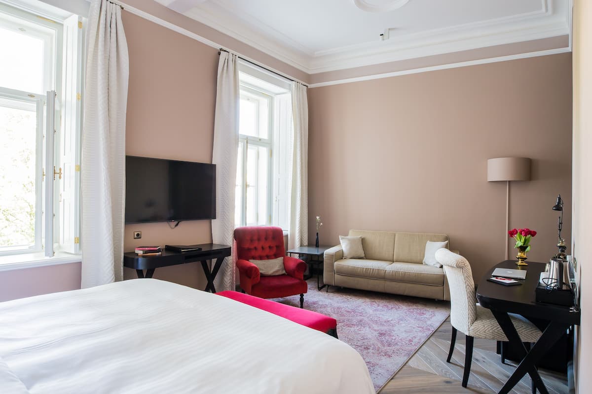 A hotel room with a large bed, a red armchair, a beige sofa, a desk with a lamp, and a TV mounted on the wall.