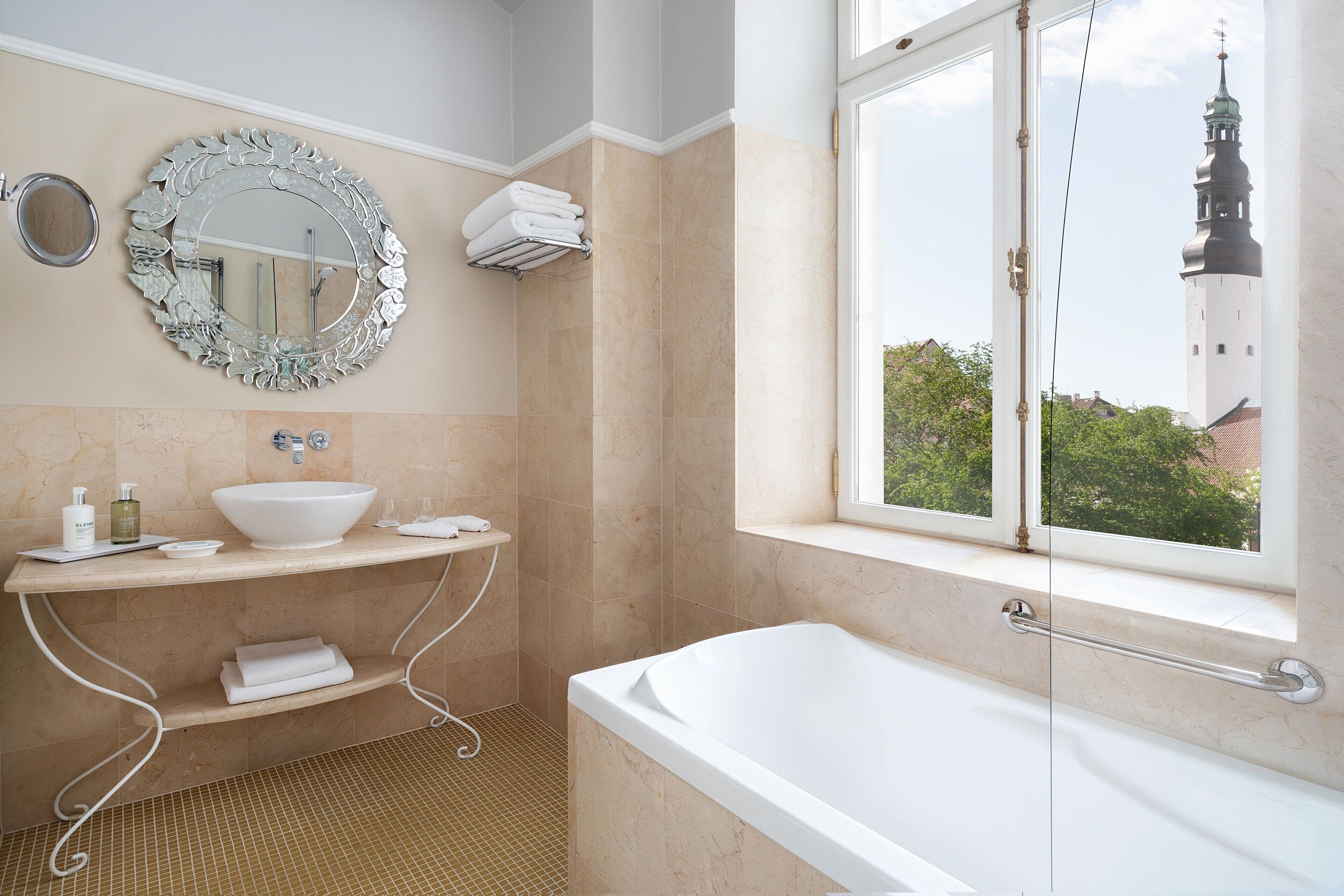 A modern bathroom with a large bathtub, a round mirror, and a window with a view of a tower.
