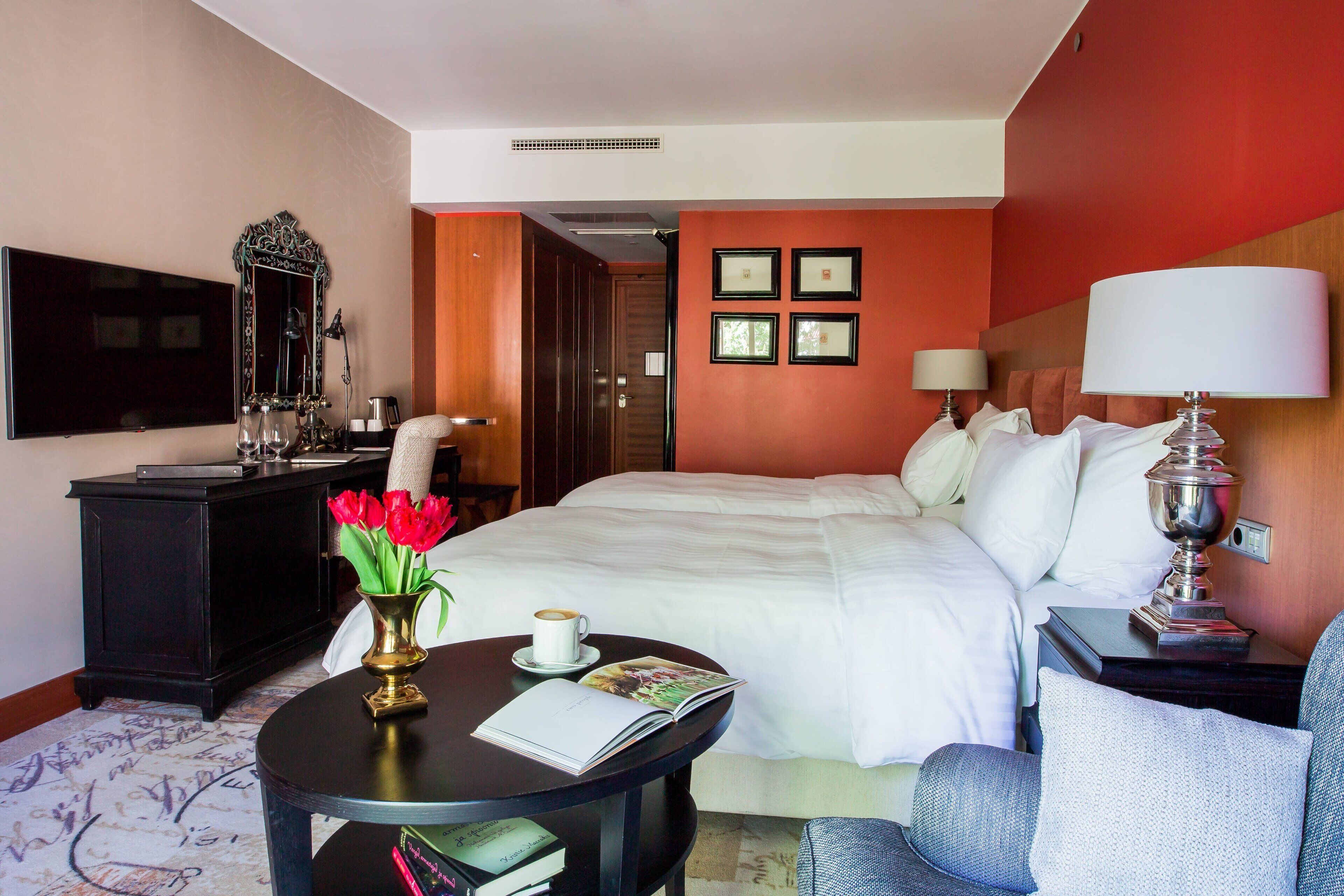 A hotel room with a bed, a desk with a lamp, a chair, a coffee table with a book and flowers, and a TV on the wall.