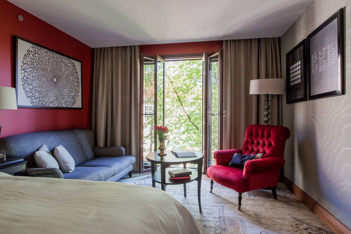 A hotel room with a bed, a red armchair, a blue sofa, a small table with a vase of flowers, and a large window with curtains.