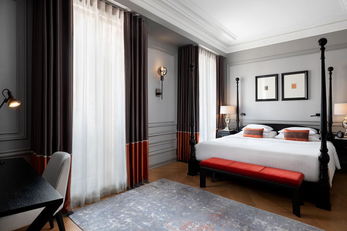 A hotel room with a large bed, a desk, and a bench. There are curtains, a wall clock, and framed pictures on the wall.