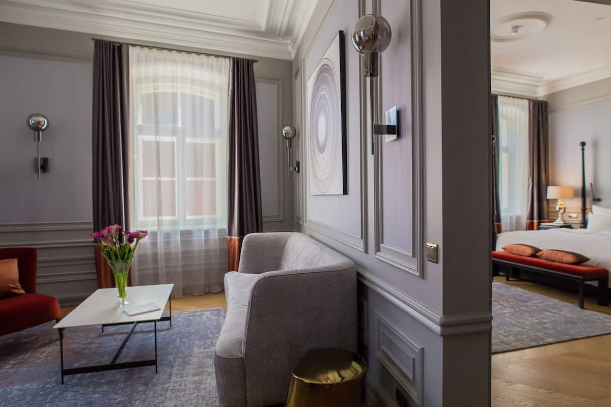 A hotel room with a grey sofa, a white coffee table, a vase with flowers, and a bed with red pillows.