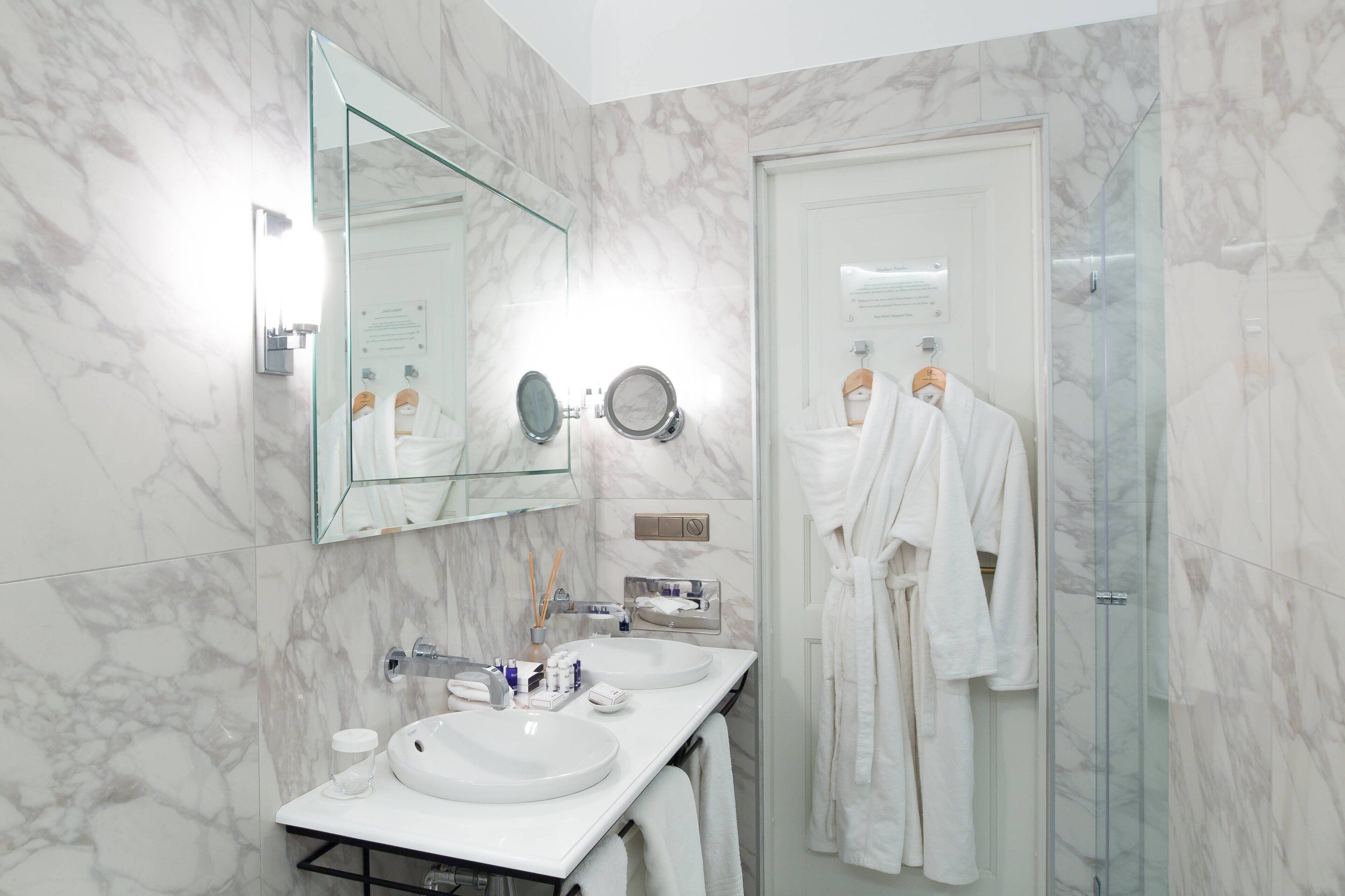 Une salle de bain moderne avec des murs en marbre, un grand miroir, deux vasques et une douche à l’italienne en verre.