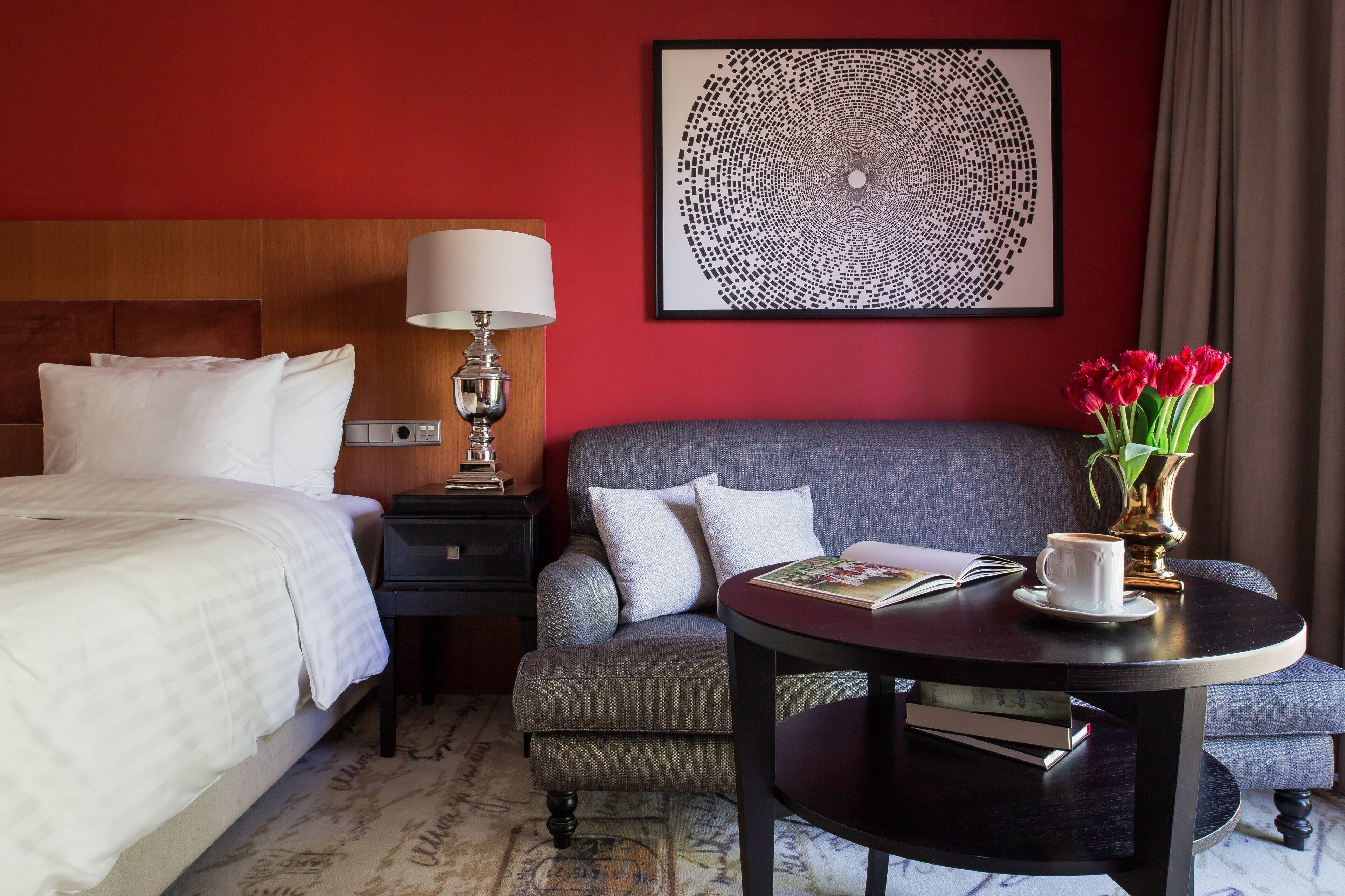 A hotel room with a bed, a grey armchair, a round table with a book and a cup, a lamp, and a framed abstract artwork on the wall.