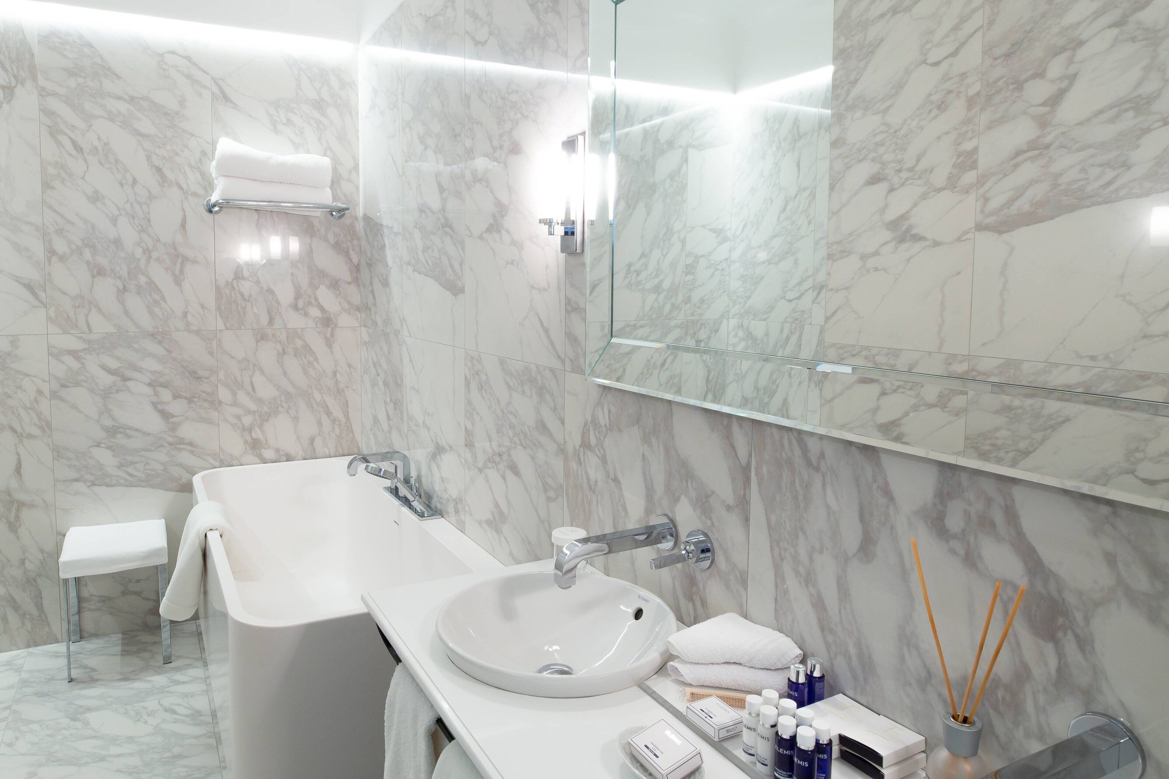 A modern bathroom with a marble wall, a white sink, a glass shower enclosure, and a small stool.