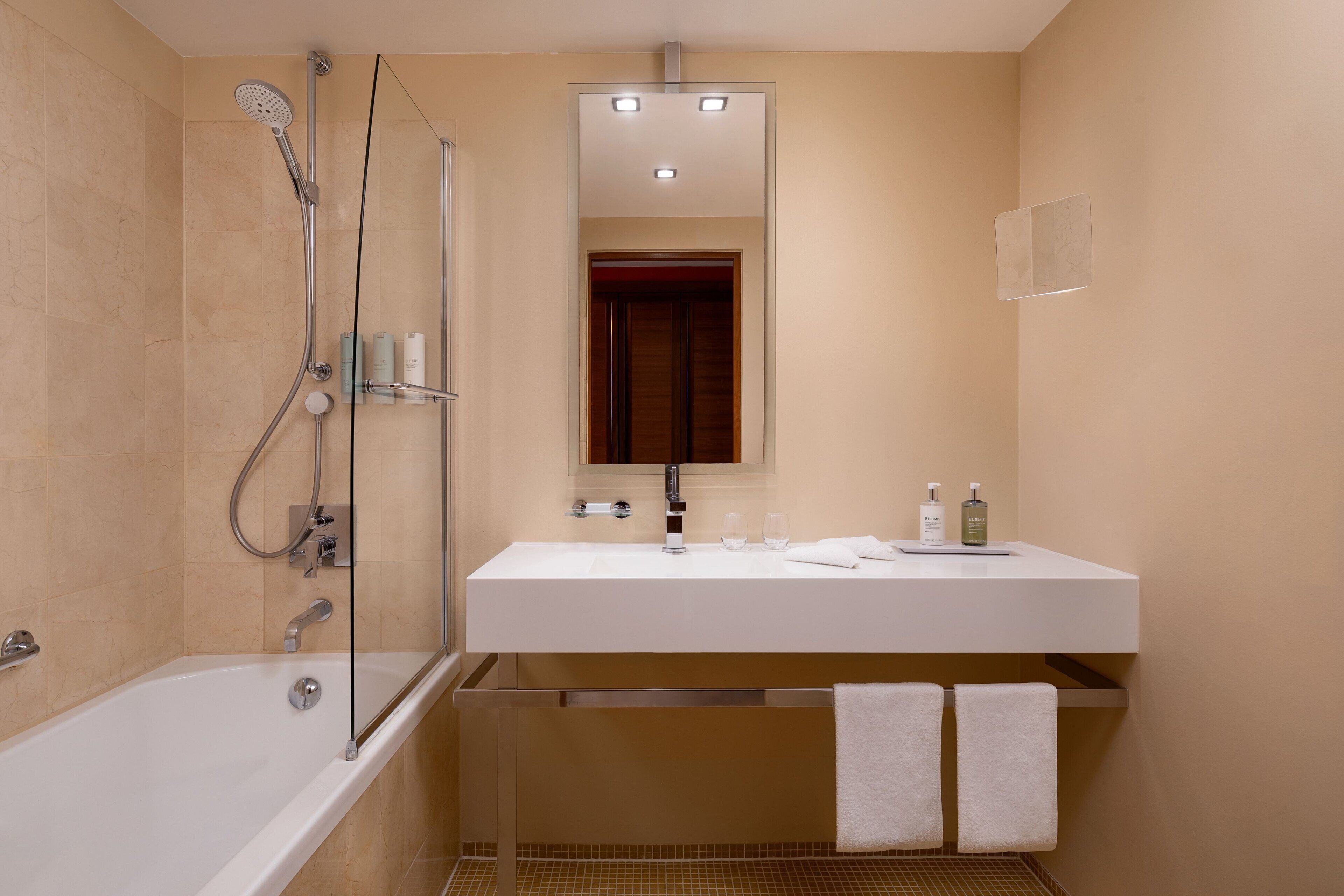 Un baño moderno con bañera, ducha, lavamanos con espejo y dos toallas colgadas en un perchero.